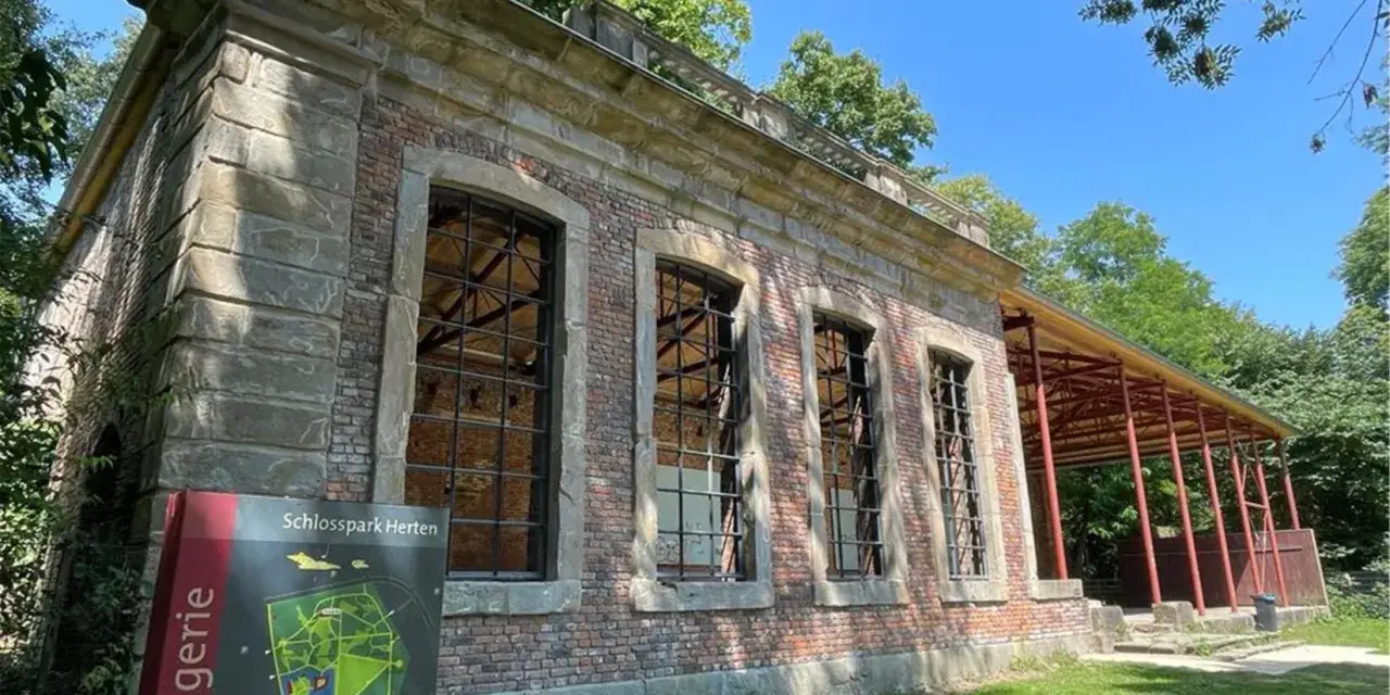 Die Orangerie Herten, ein historisches Geb&auml;ude mit gro&szlig;en Fensterfronten und einem roten Vordach, steht im Schatten von B&auml;umen.