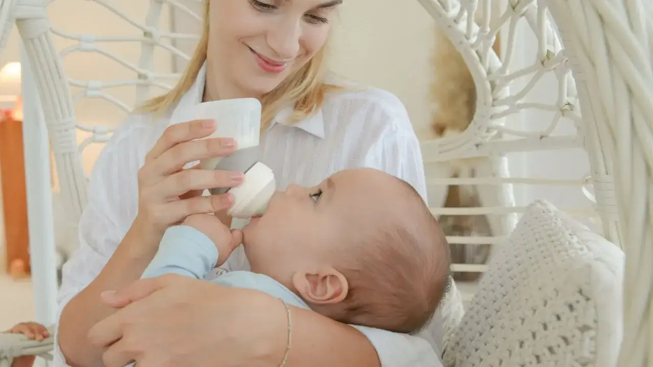 A mother smiles while holding her baby for upright bottle feeding. The baby is looking up at her, taking milk from the bottle.