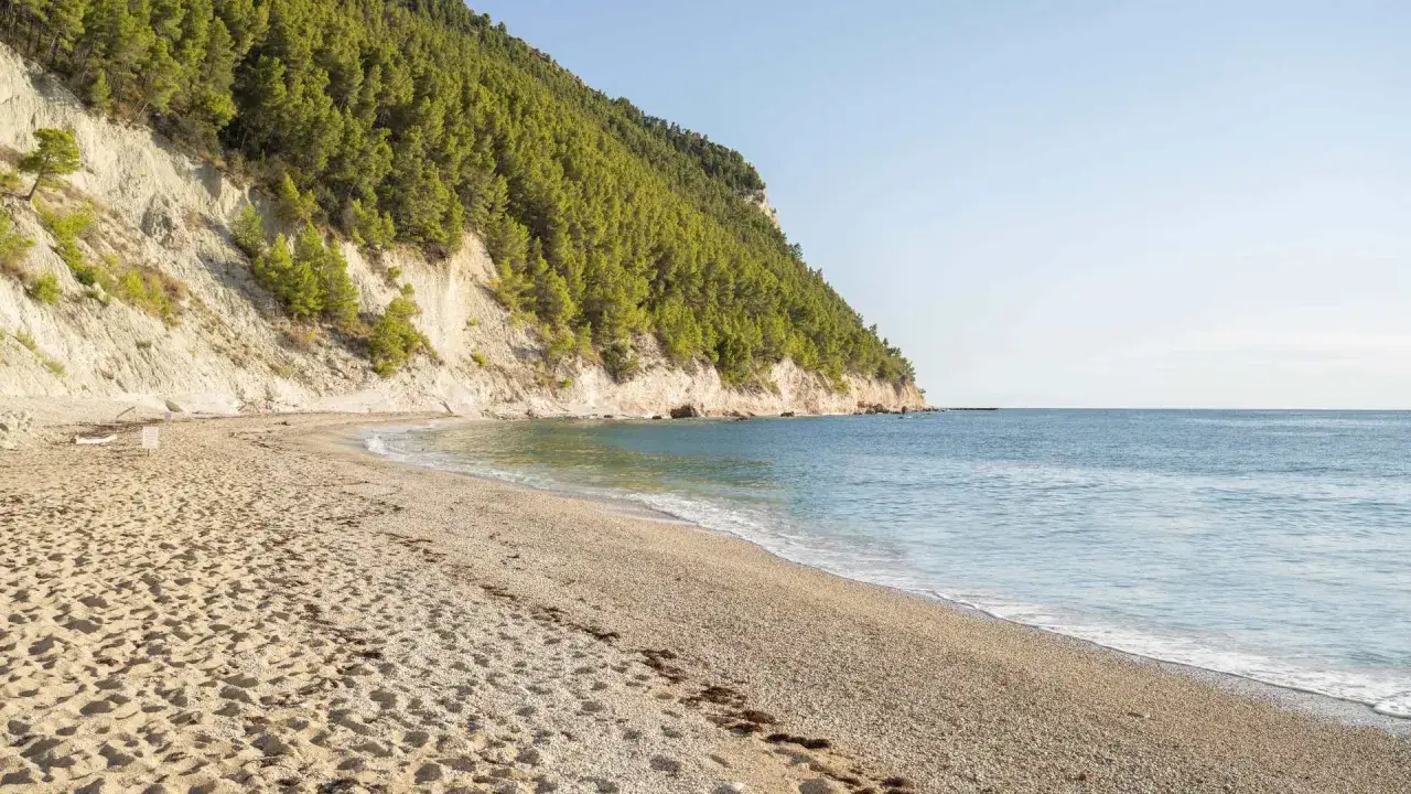 La **spiaggia** di **Castelmezzano** si estende serena, con sabbia dorata e ciottoli levigati, lambita da un mare azzurro e calmo sotto una scogliera verdeggiante.