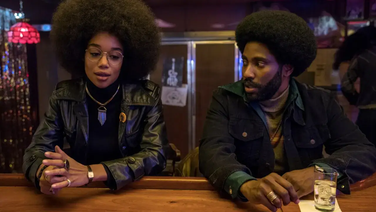 Ron och Patrice i Blackkklansman, med sina ikoniska frisyrer och kl&auml;der, sitter vid en bar och diskuterar.