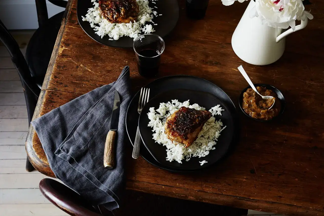 Tv&aring; tallrikar med ris och k&ouml;tt, en sk&aring;l med rabarber chutney och en vas med blommor p&aring; ett tr&auml;bord.