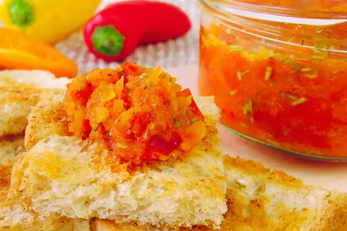 Fette di pane tostato con una generosa porzione di crema di peperoni, accanto a un barattolo pieno della stessa prelibatezza. Peperoni rossi, gialli e arancioni sullo sfondo.