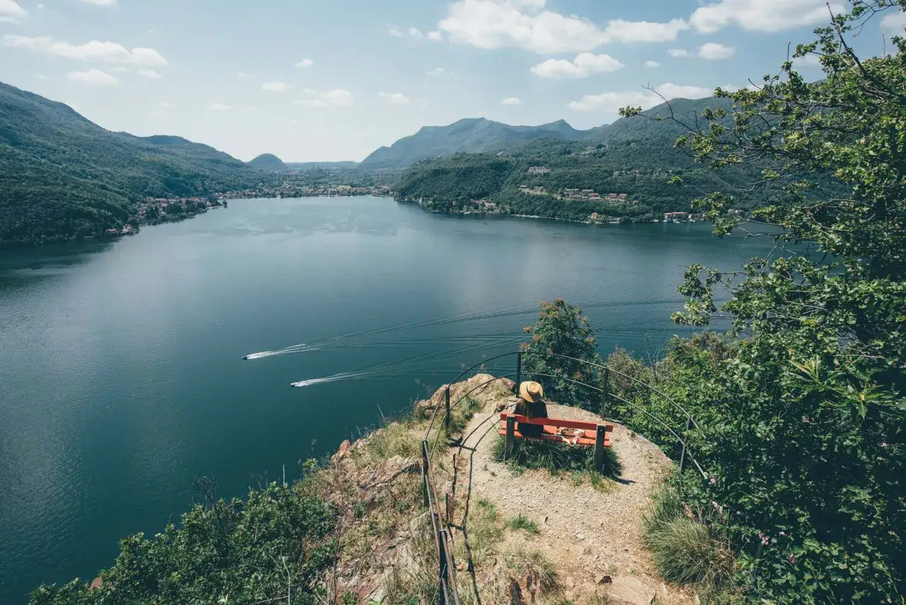 Une personne assise sur un banc admire le lac de Lugano et les montagnes verdoyantes. Deux bateaux filent sur l'eau.