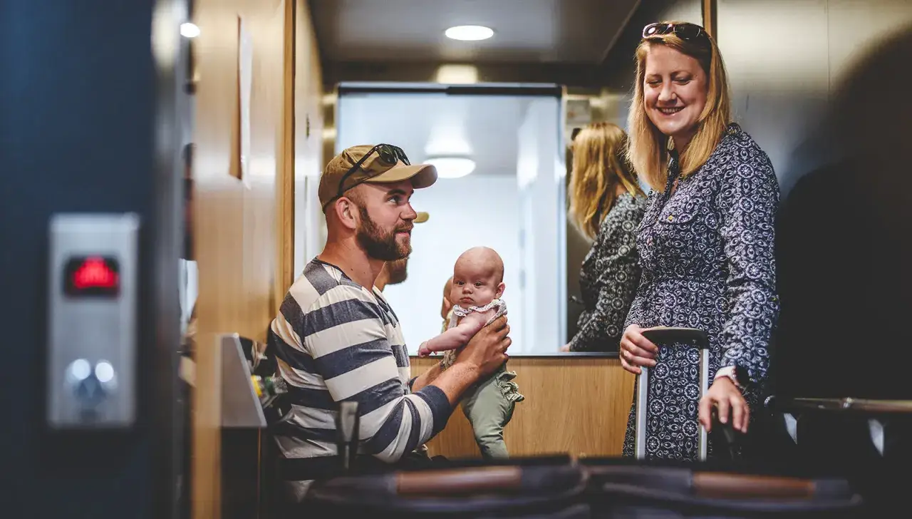 Familj i en gammal hiss. Pappa håller sitt barn, mamma står bredvid med resväska.
