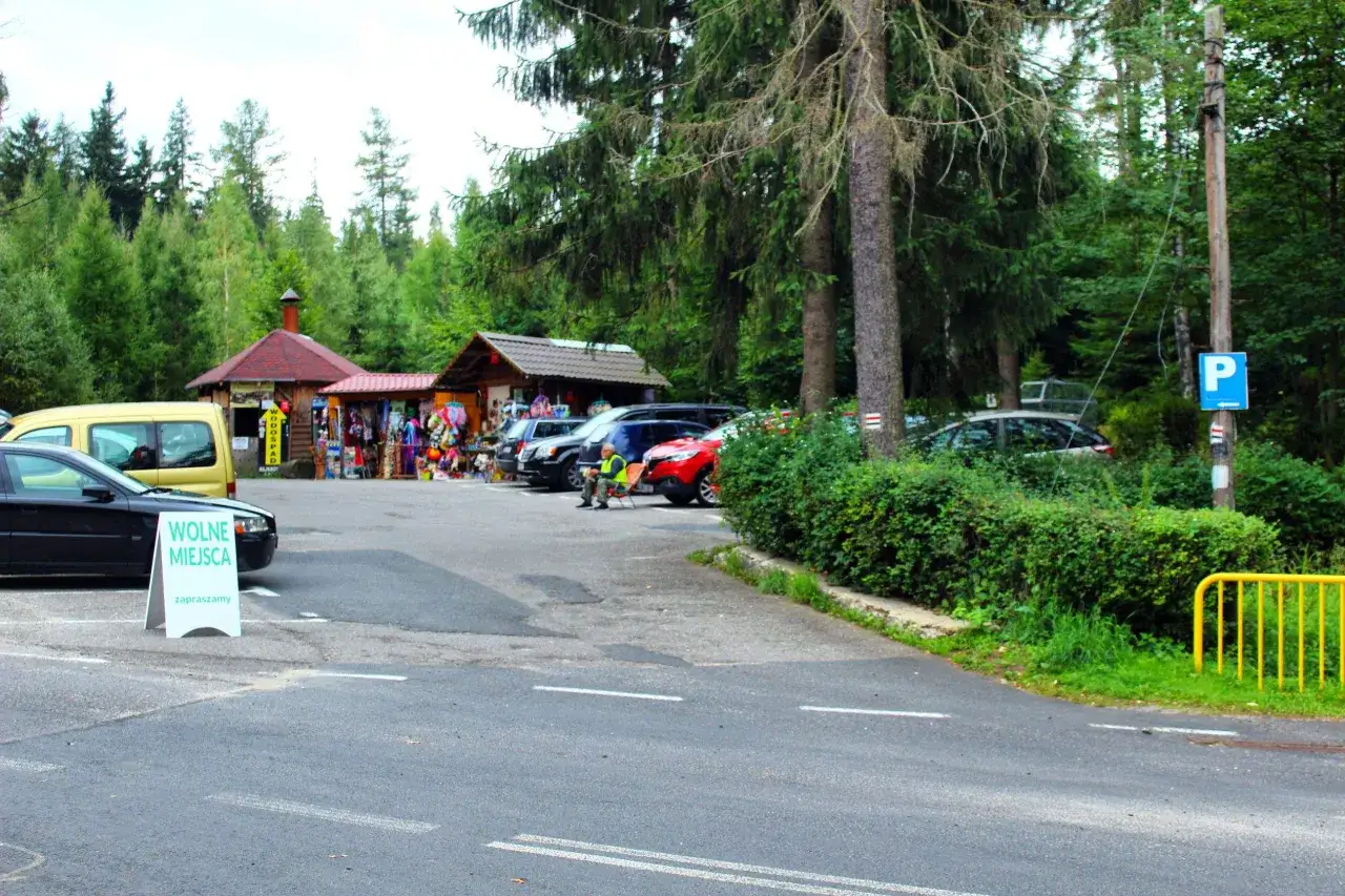Parking przy Wodospadzie Kamieńczyka, najkrótsza trasa. Stoiska z pamiątkami, samochody i zielone drzewa.