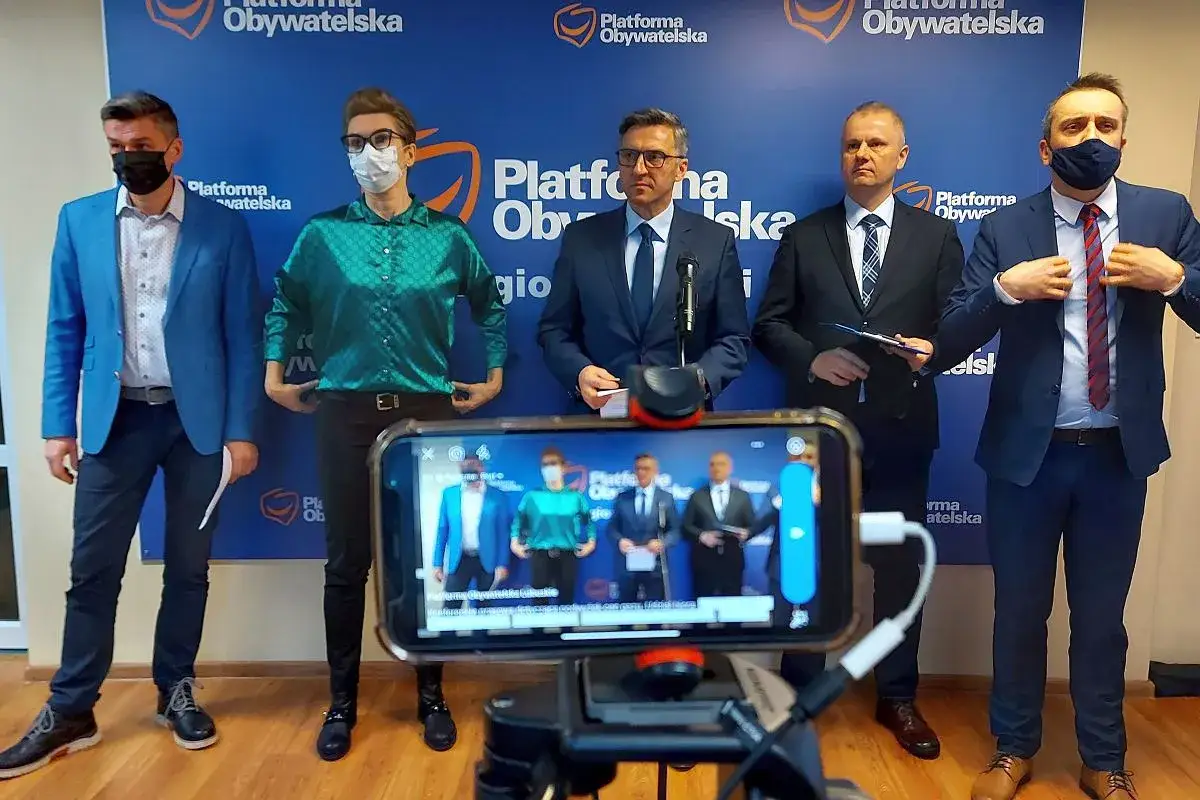 Pięciu mężczyzn w garniturach stoi przed niebieskim banerem z logo Platformy Obywatelskiej. Na pierwszym planie widać ekran telefonu z transmisją na żywo.