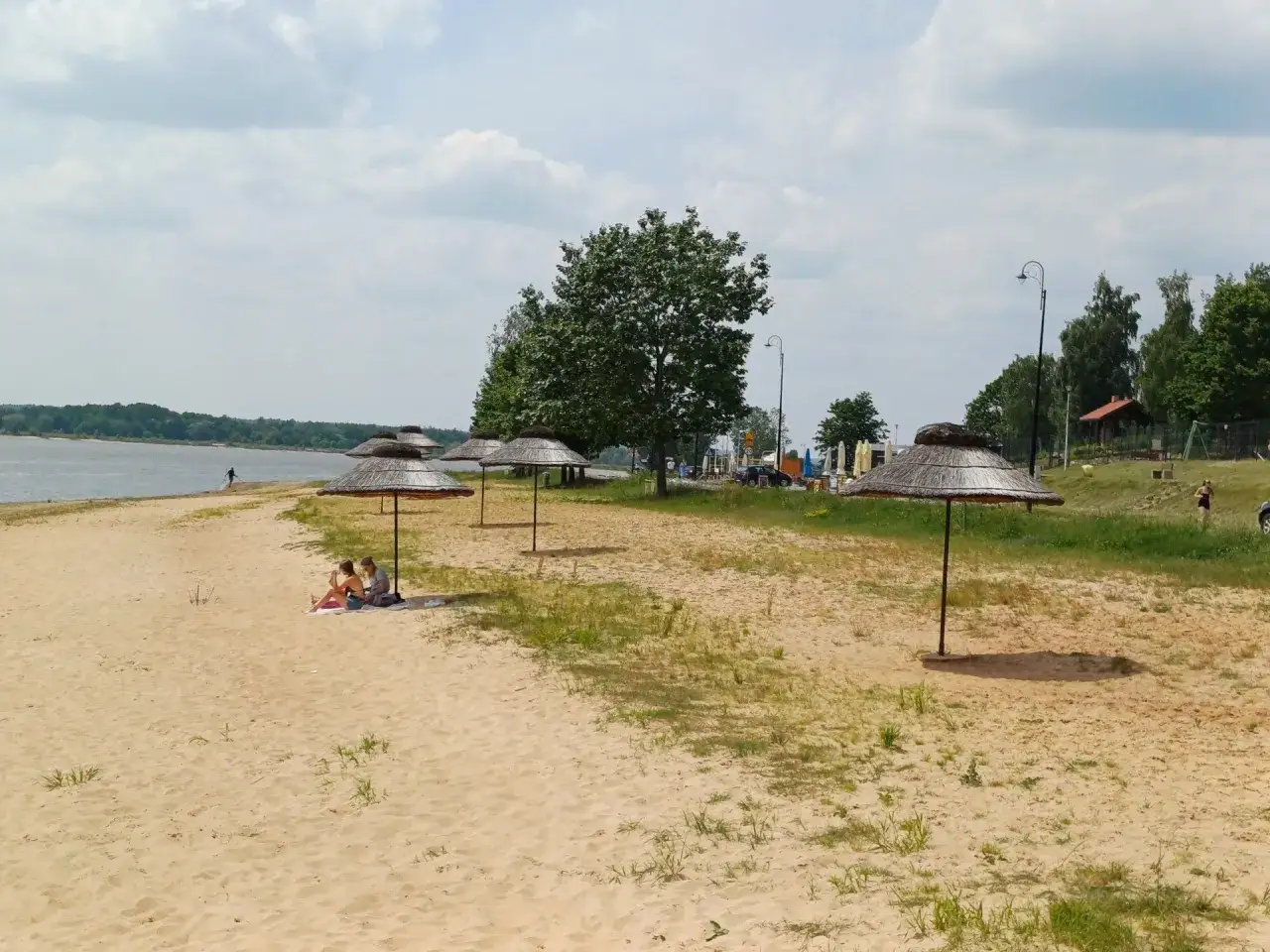 Słoneczny dzień nad zalewem Brody. Piasek, parasole i kilka osób odpoczywających na plaży.
