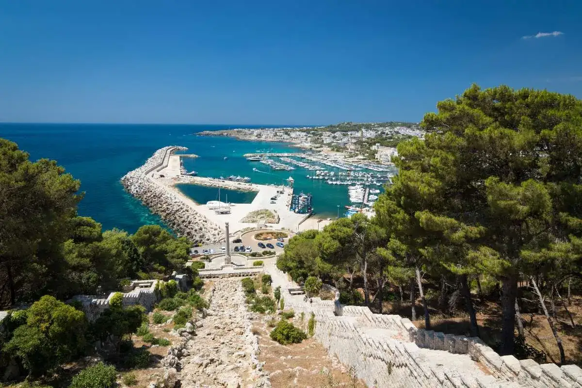 Un suggestivo cammino del Salento conduce a un porto con barche, mare azzurro e un borgo bianco.
