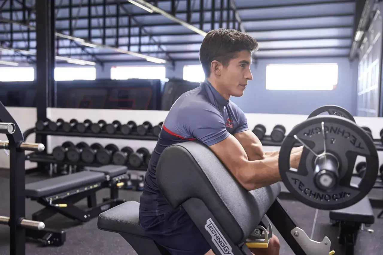 Marc Márquez, con su físico atlético, entrena bíceps en el gimnasio con una barra.