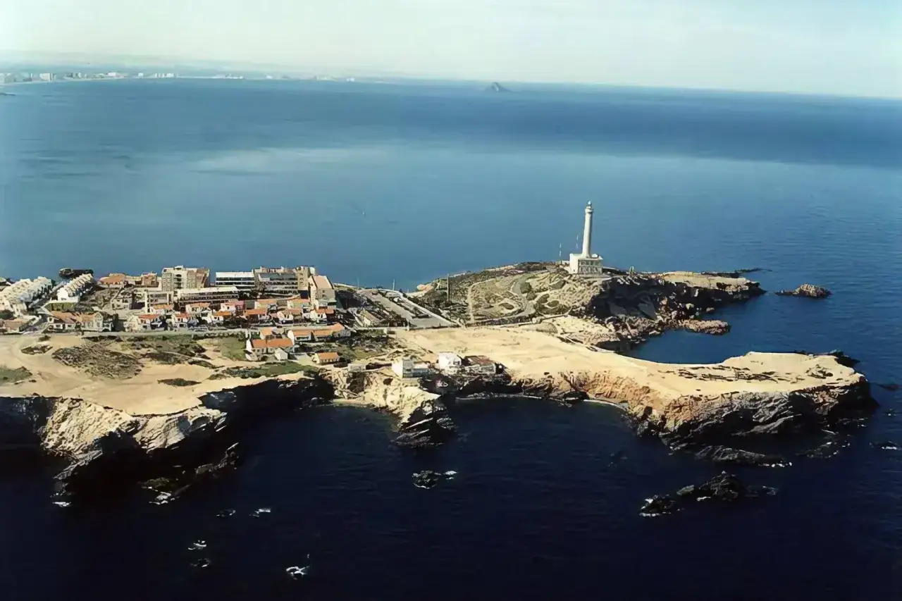 Vista aérea de un pueblo costero con un faro blanco en un promontorio rocoso. El mar azul profundo rodea la **cala roja** y las casas blancas.