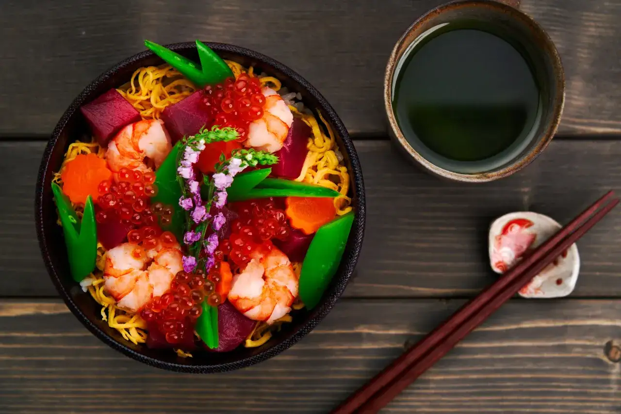 Bunte chirashi bowl mit Garnelen, Thunfisch, Lachsrogen und Gem&uuml;se, daneben eine Tasse gr&uuml;nen Tees.