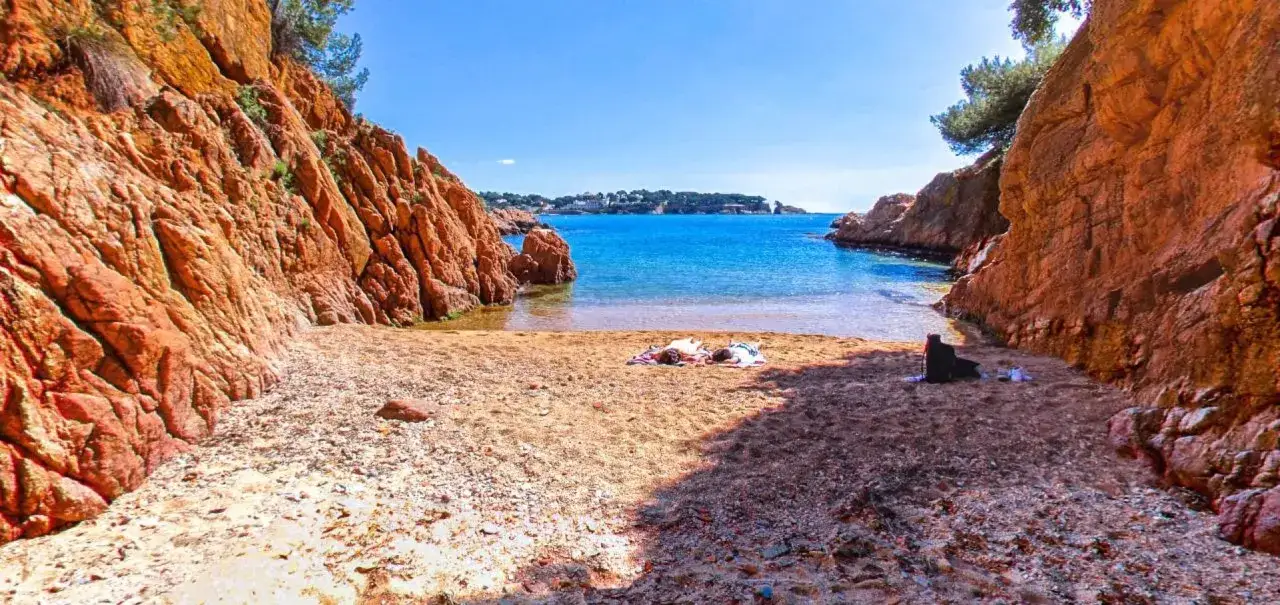 Dos personas toman el sol en la arena dorada de cala Maset, rodeada de acantilados rojizos y aguas turquesas.