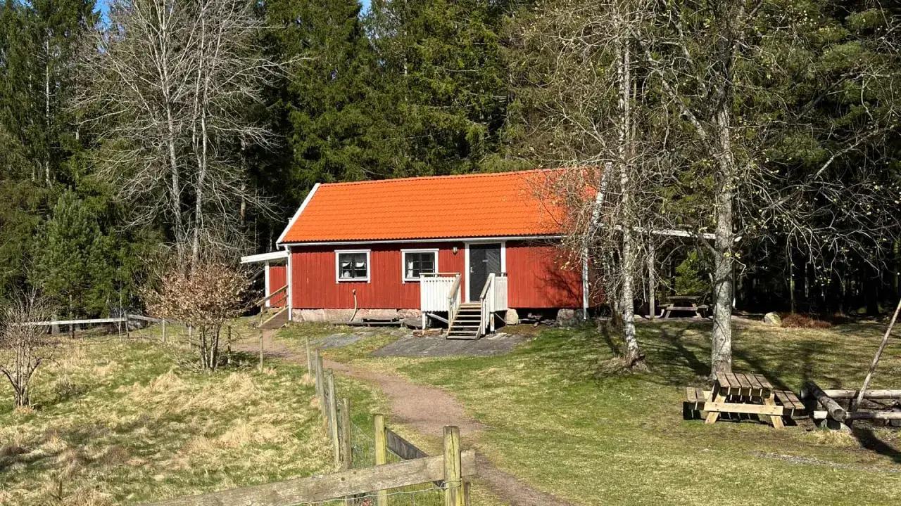 Rött torp med orange tak, omgivet av skog. En stig leder fram till torpet Freden, där picknickbord står utplacerade.