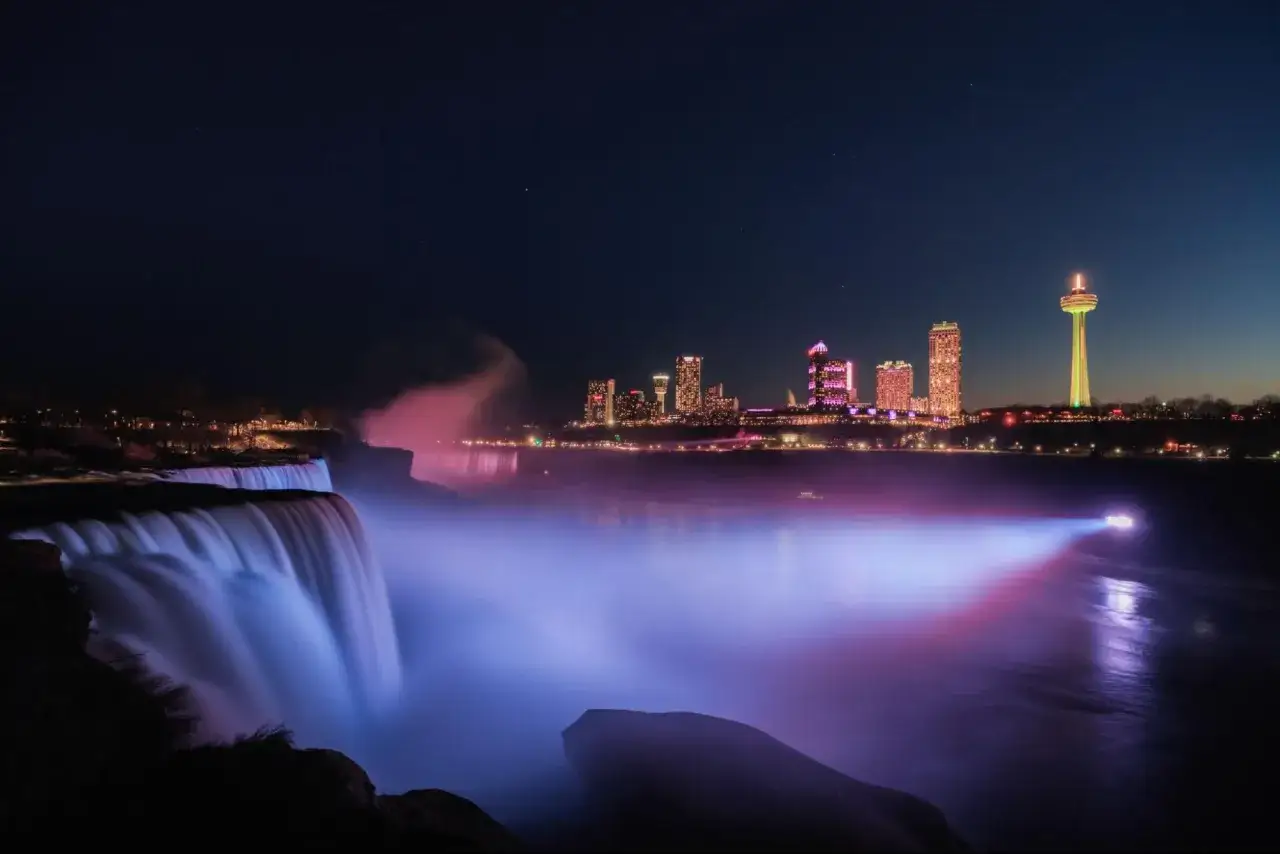 Las cataratas del Ni&aacute;gara iluminadas de noche, con la silueta de la ciudad y una torre alta.