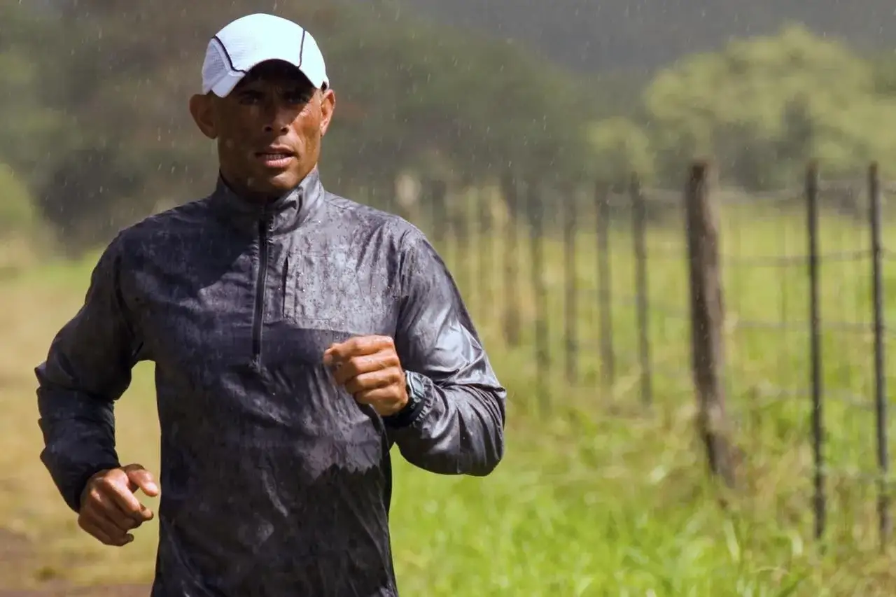 Uomo con cappellino bianco corre sotto la pioggia battente, la sua giacca &egrave; bagnata.
