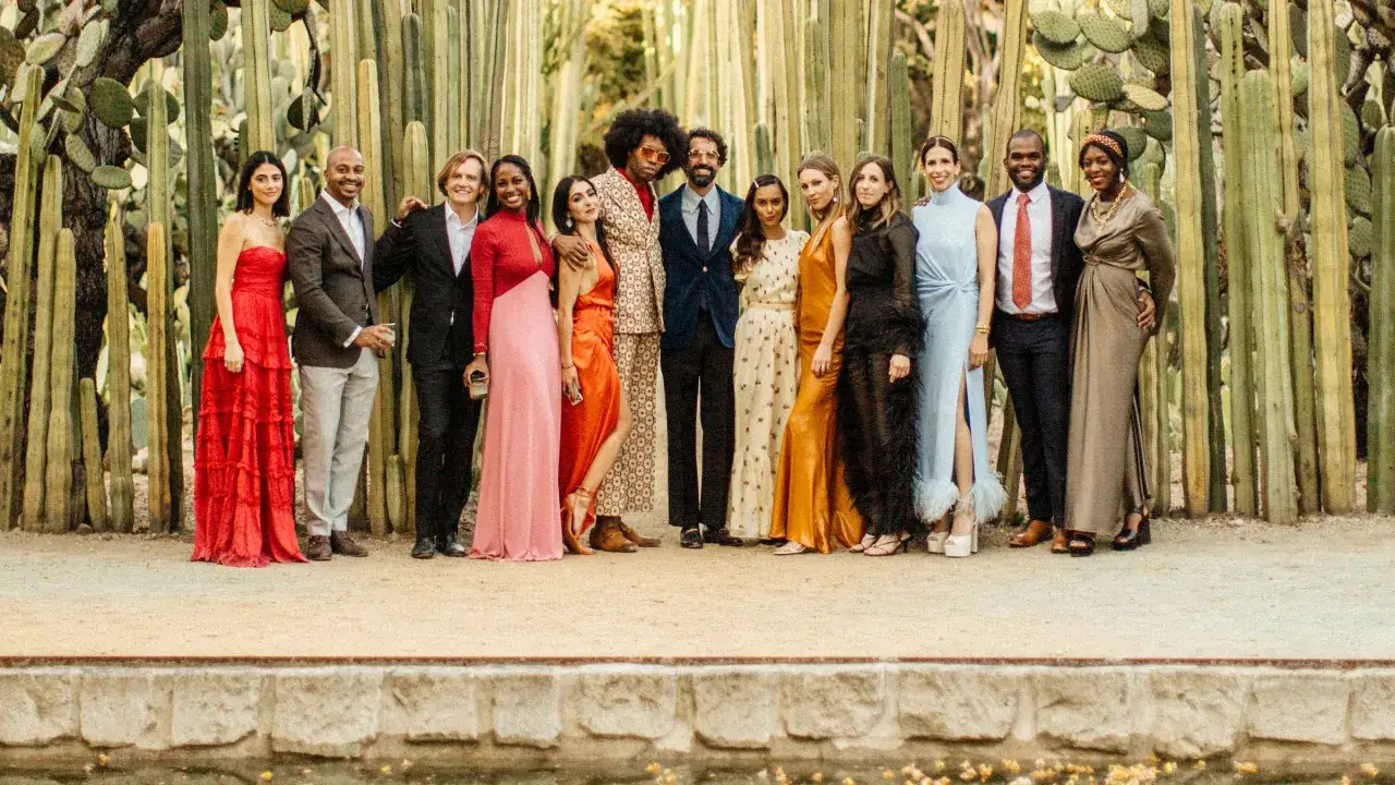 Un gruppo di persone eleganti, alcune con abiti da sera e altre con completi, posa davanti a cactus. Un uomo indossa un completo con **black tie**.