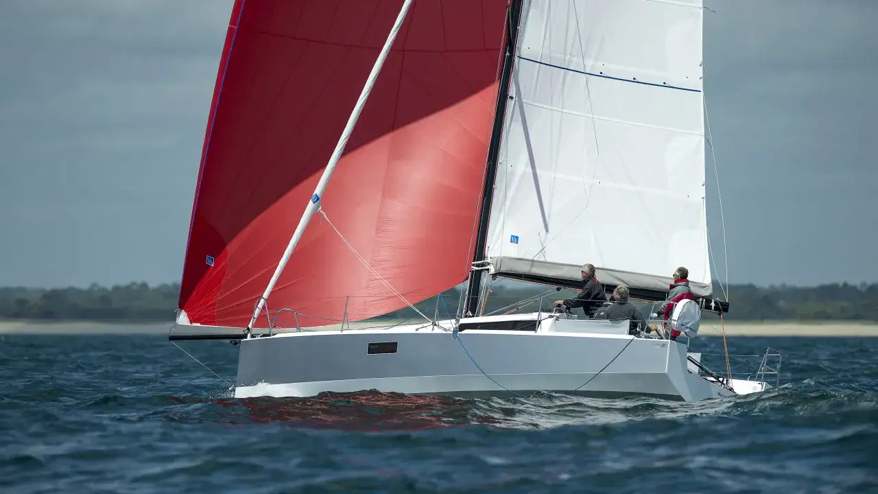 Un voilier Pogo 30 navigue sur une mer agitée, ses voiles rouge et blanche gonflées par le vent. Les avis sur ce bateau sont positifs.