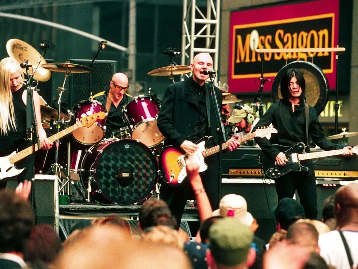 Les membres de The Smashing Pumpkins jouent sur sc&egrave;ne. Une guitariste blonde, un batteur chauve, et deux guitaristes chauves et aux cheveux noirs.