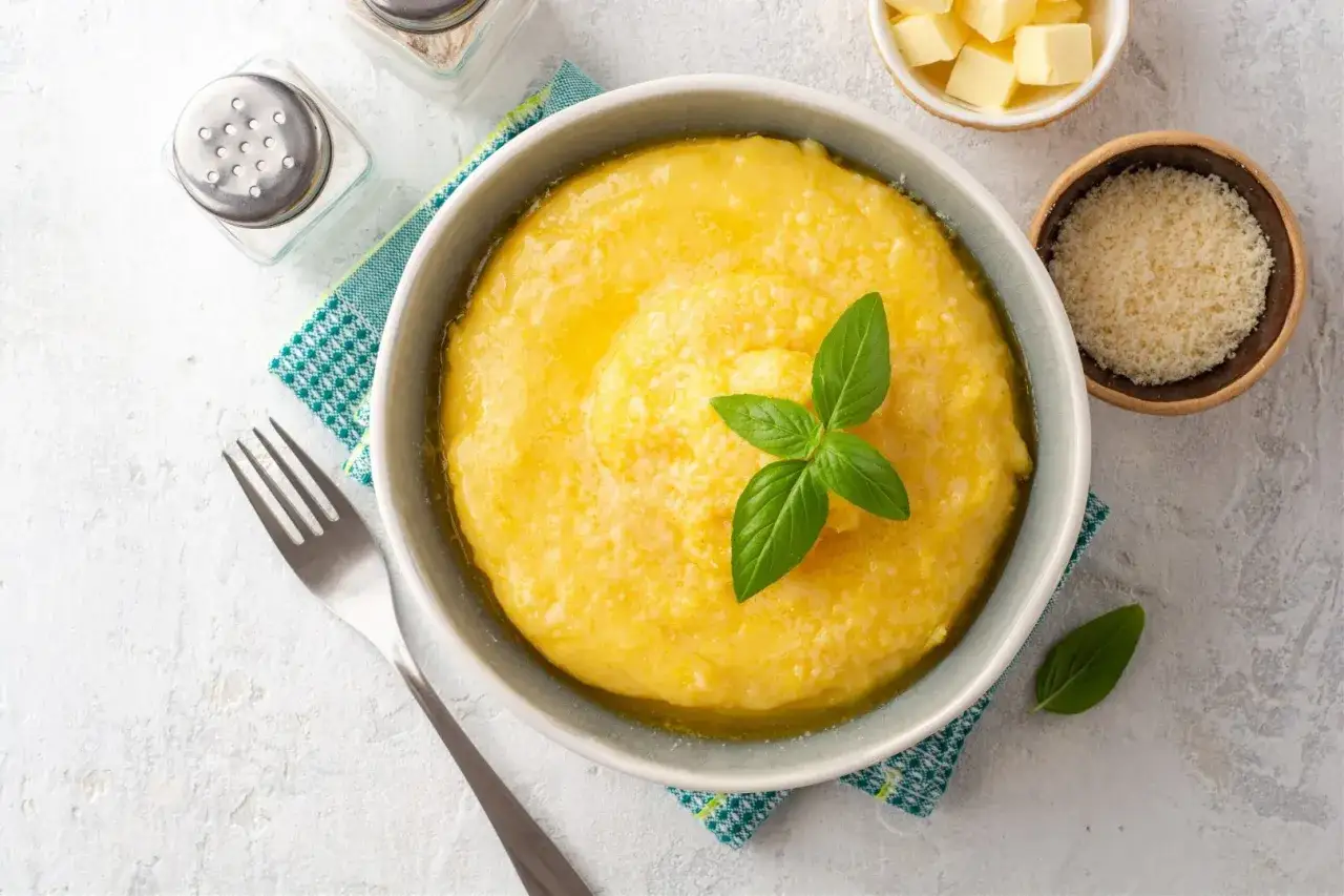 A miseczce kremowa polenta, udekorowana listkami bazylii, obok miseczki z parmezanem i kostkami masła.