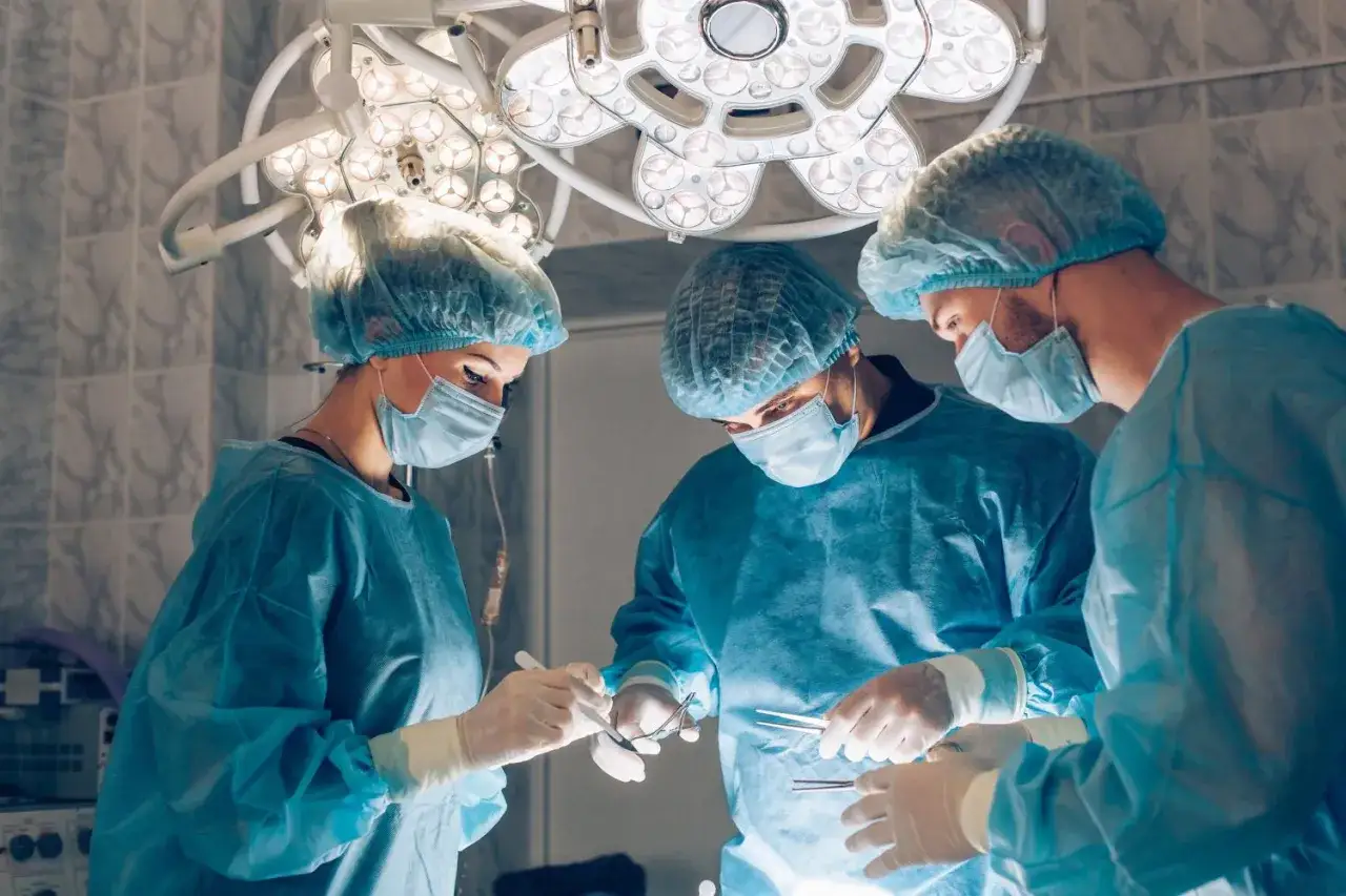 Zespół chirurgów w maseczkach i czepkach wykonuje skomplikowany zabieg na barku pod jasnym światłem lampy operacyjnej.