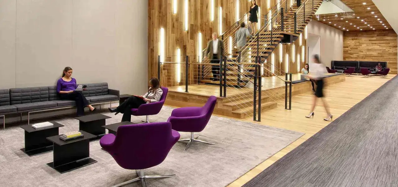 Moderna sala di attesa con poltrone viola, divano grigio e una scala in legno illuminata da luci verticali.