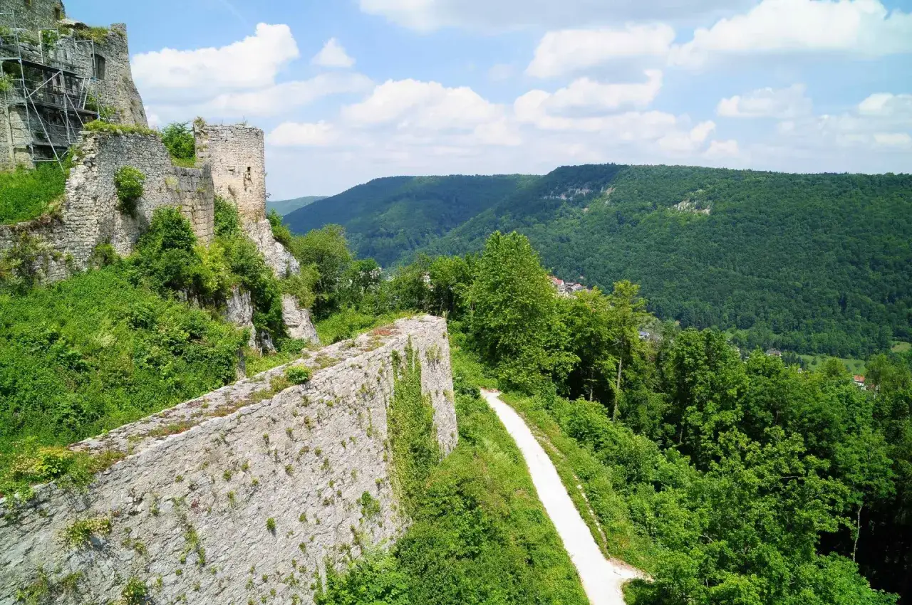 Die Ruine Hofen thront auf einem Felsen, umgeben von &uuml;ppigem Gr&uuml;n. Ein schmaler Pfad f&uuml;hrt entlang der alten Mauern, w&auml;hrend sich im Hintergrund bewaldete H&uuml;gel erstrecken.