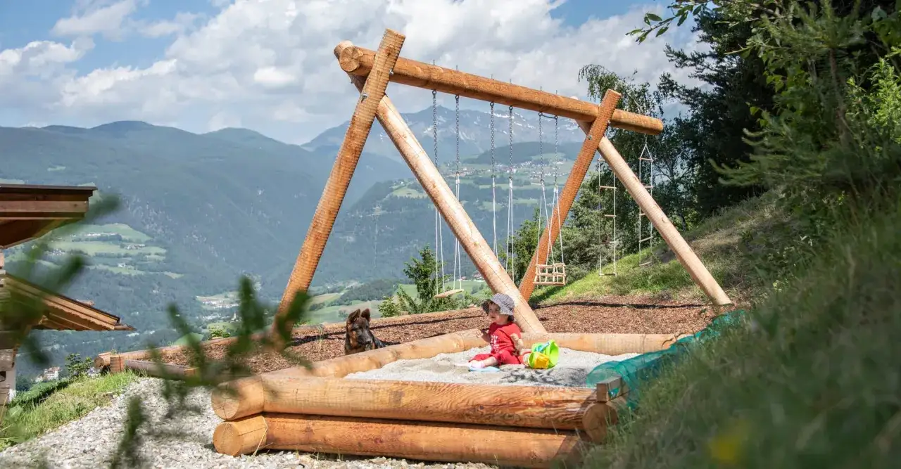 Bambino gioca nella sabbiera con un cane, sullo sfondo le fattorie altoatesine e montagne verdi.