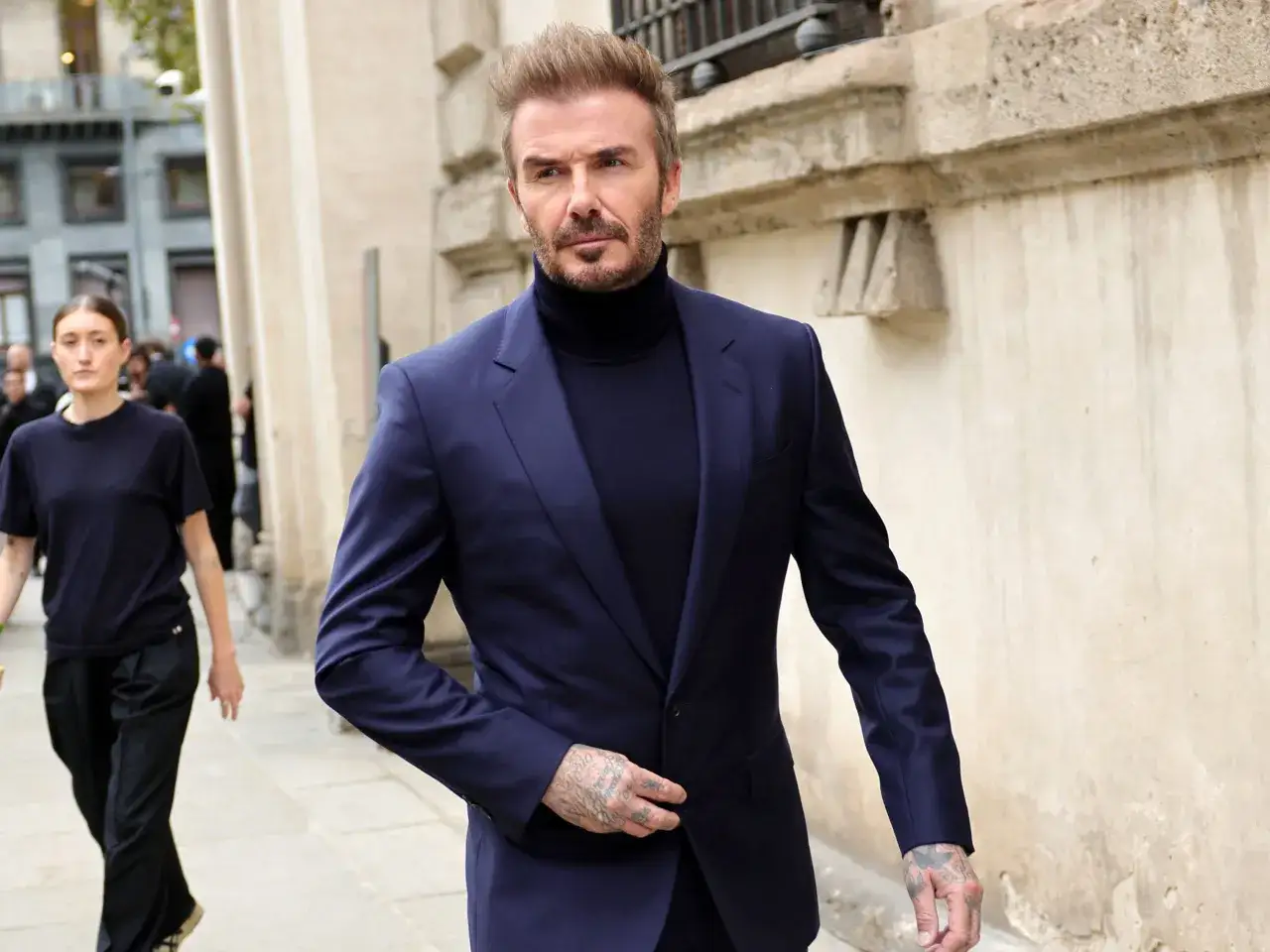 David Beckham luce un elegante traje azul con jersey de cuello alto, caminando con confianza.