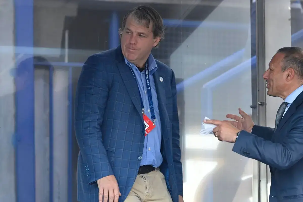 Todd Boehly, właściciel Chelsea, w niebieskiej marynarce i z identyfikatorem UEFA Champions League, rozmawia z mężczyzną w garniturze.