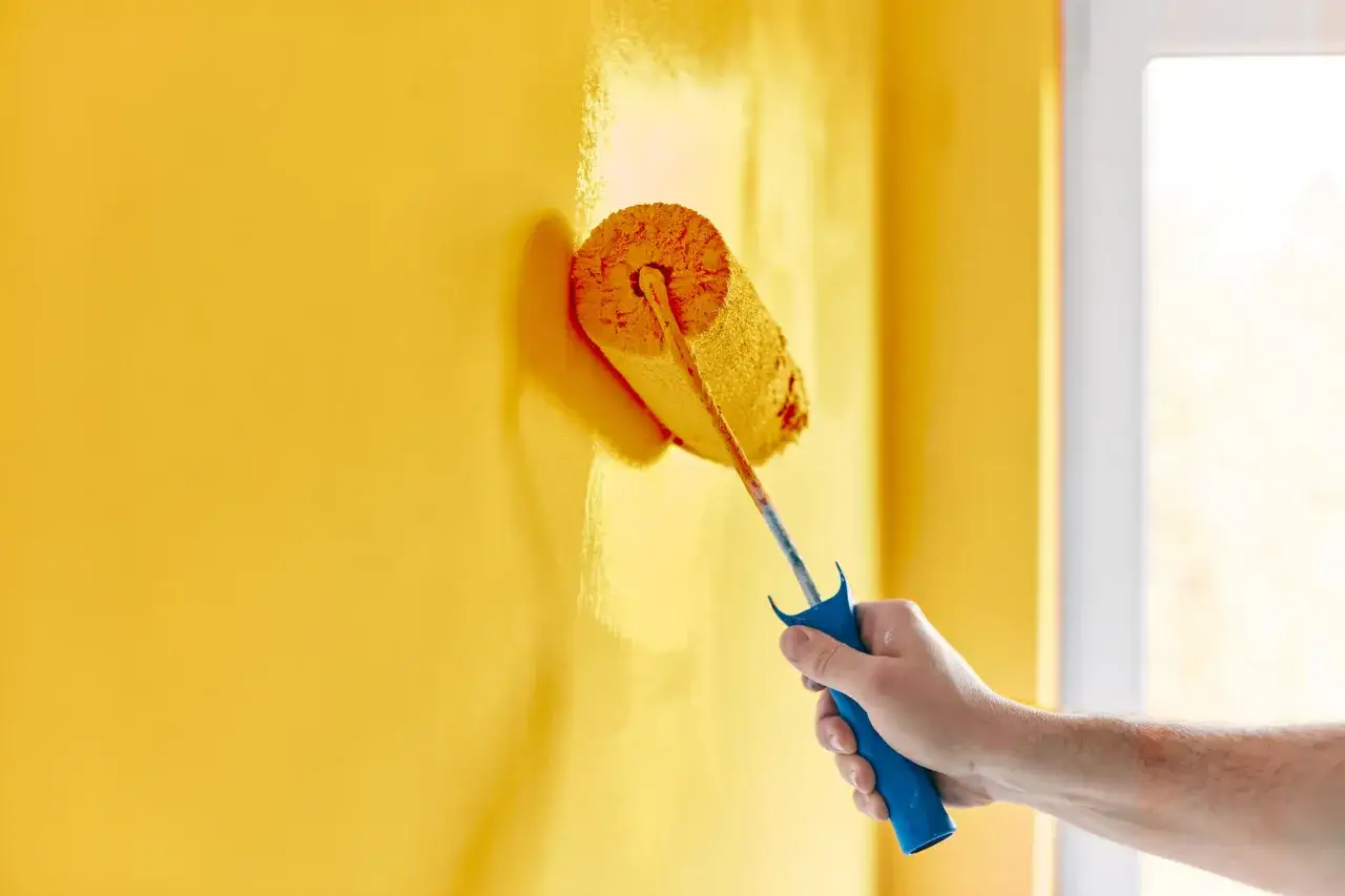 Mano che imbianca una parete gialla con un rullo. Il periodo migliore per imbiancare casa &egrave; quando c'&egrave; buona ventilazione.