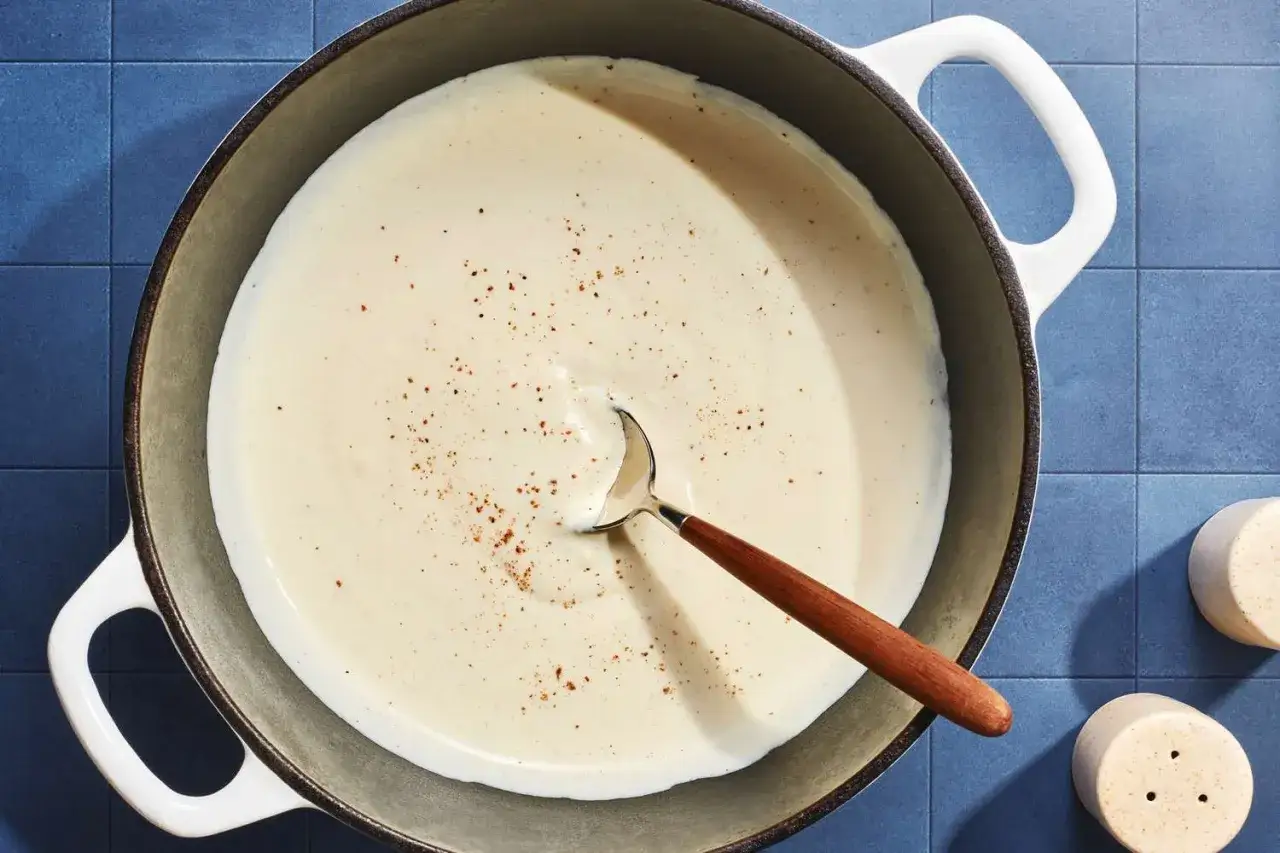 A creamy sauce, likely crème fraîche, simmers in a pot, seasoned with pepper. A wooden-handled spoon rests in the rich mixture.