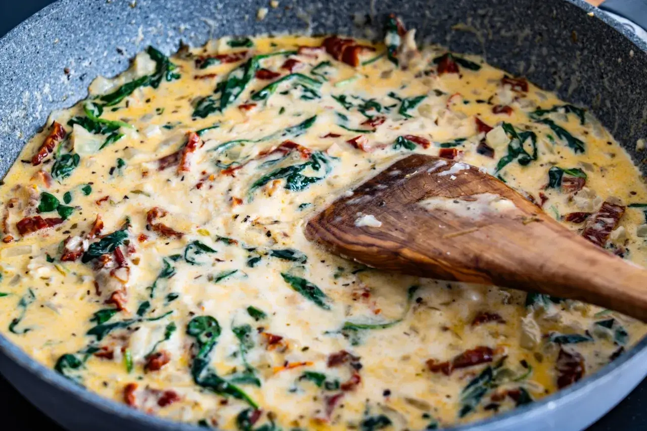 Une po&ecirc;le de p&acirc;tes &agrave; la toscane cr&eacute;meuses avec des &eacute;pinards et des tomates s&eacute;ch&eacute;es, m&eacute;lang&eacute;es avec une cuill&egrave;re en bois.