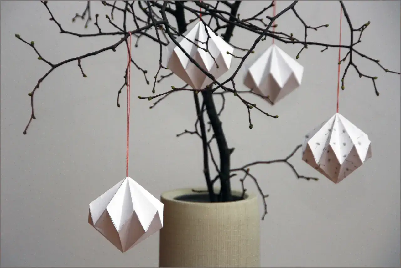 Origami-Diamanten hängen an kahlen Zweigen. Eine Vorlage zum Diamant basteln könnte diese filigranen Dekorationen inspirieren.