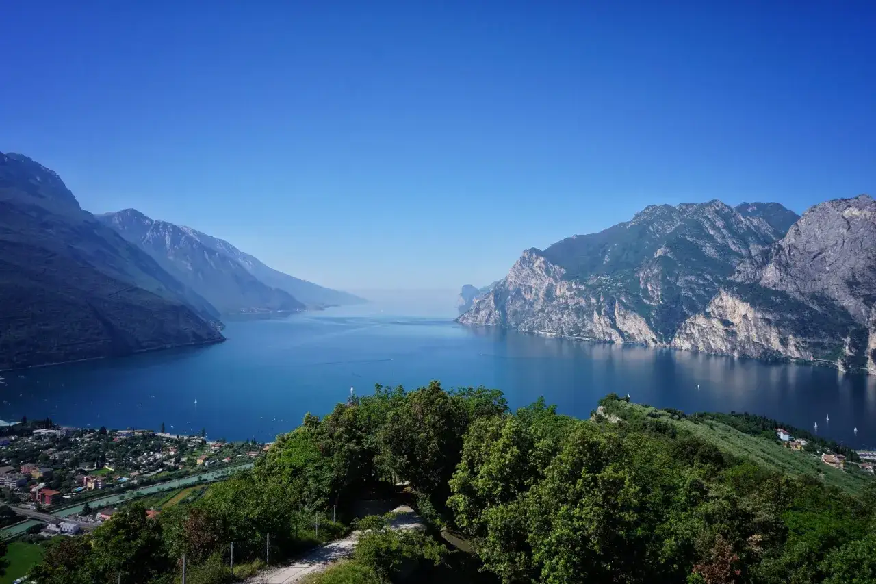 Blick von oben auf den Gardasee mit majestätischen Bergen, die sich im tiefblauen Wasser spiegeln. Kleine Boote gleiten über die ruhige Oberfläche.