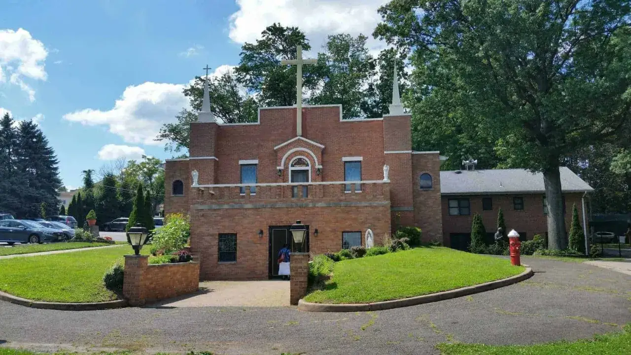 The Holy Face Monastery, a brick building with a large cross, stands under a blue sky with fluffy clouds.