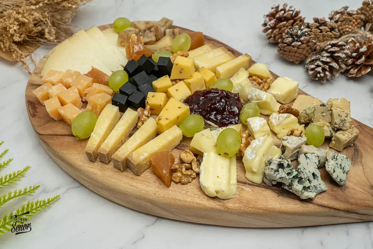 Una exquisita presentaci&oacute;n tabla de quesos con variedad de texturas y sabores, uvas, nueces y mermelada.