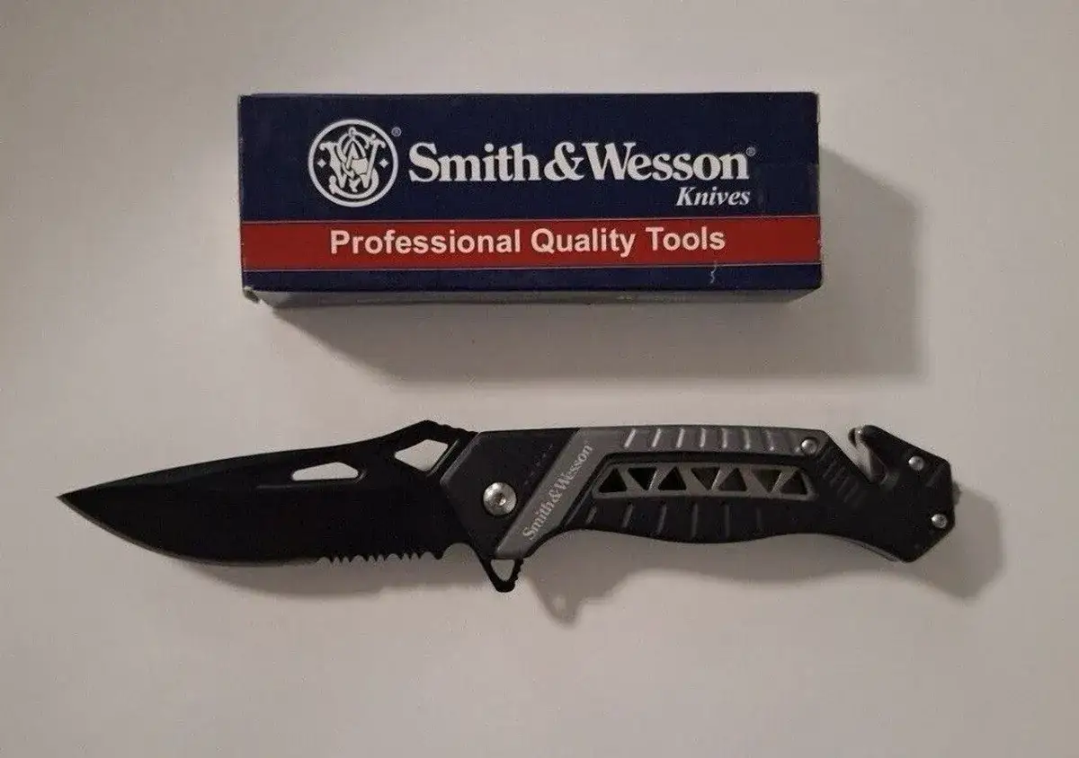 Navaja Smith & Wesson negra con hoja dentada y mango ergon&oacute;mico, junto a su caja 