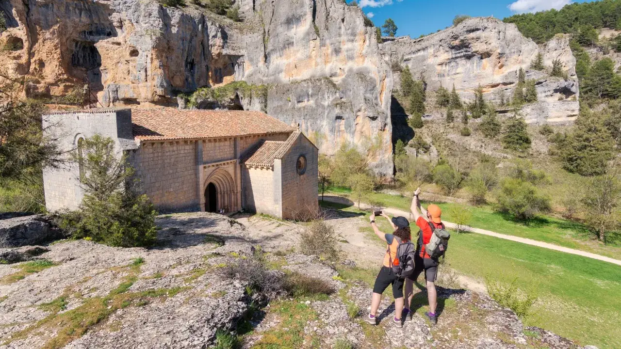 Pareja en la **ruta de las ermitas**, fotografiando una iglesia rom&aacute;nica junto a un imponente farall&oacute;n rocoso.