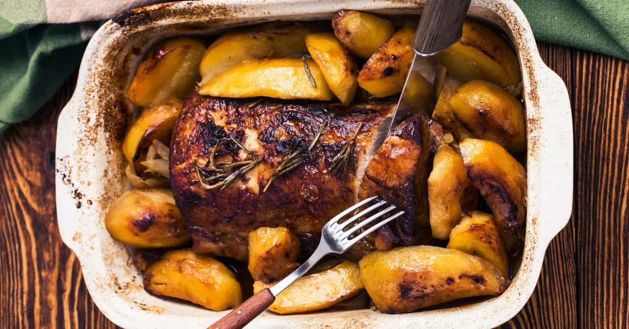 Schab pieczony z ziemniakami w naczyniu, skropiony ziołami. Nóż i widelec gotowe do krojenia.
