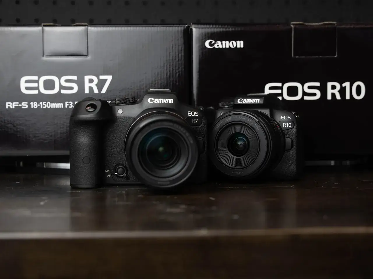 Due fotocamere Canon EOS R7 e R10, entrambe con sensore APS-C, affiancate su un tavolo scuro, con le loro scatole sullo sfondo.