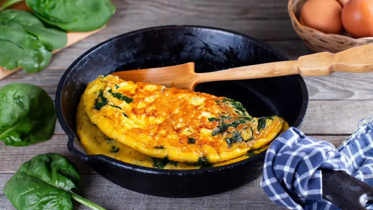 Une omelette simple aux &eacute;pinards, dor&eacute;e et moelleuse, servie dans une po&ecirc;le en fonte. Des feuilles d'&eacute;pinards frais et des &oelig;ufs sont dispos&eacute;s autour.