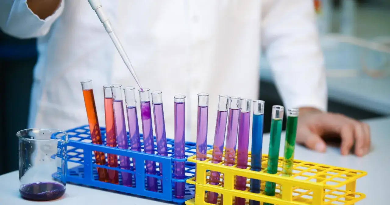 Laborantka dodaje płyn do probówki, badając potencjalną kwasicę. Kolorowe płyny w probówkach sugerują różne reakcje chemiczne.