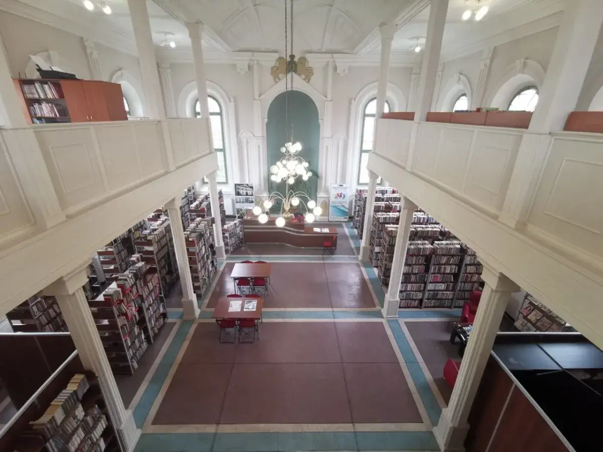 Wnętrze biblioteki w Sompolnie, z regałami pełnymi książek na dwóch poziomach, stołami do czytania i ozdobnym żyrandolem.