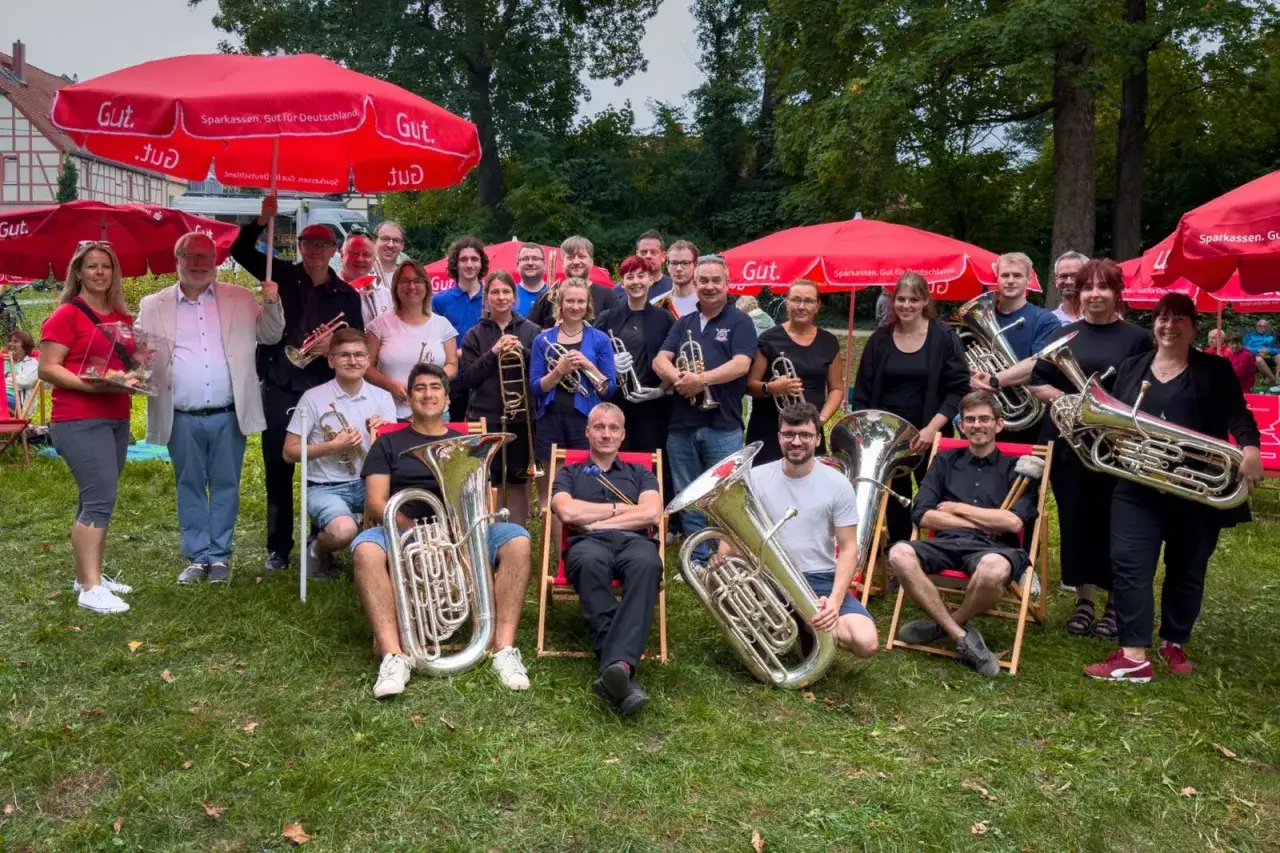Eine fröhliche Brass Band posiert unter roten Sonnenschirmen. Viele Mitglieder halten ihre Instrumente, bereit für ein Konzert.