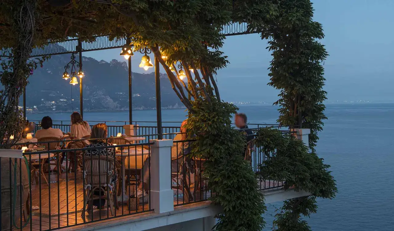 Persone gustano un caff&egrave; al mare, seduti in un pergolato illuminato da lanterne, con vista su montagne e mare al crepuscolo.