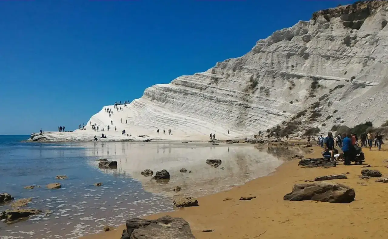 Sycylia najpiękniejsze miejsca: białe klify Scala dei Turchi odbijają się w spokojnym morzu, a turyści spacerują po plaży.