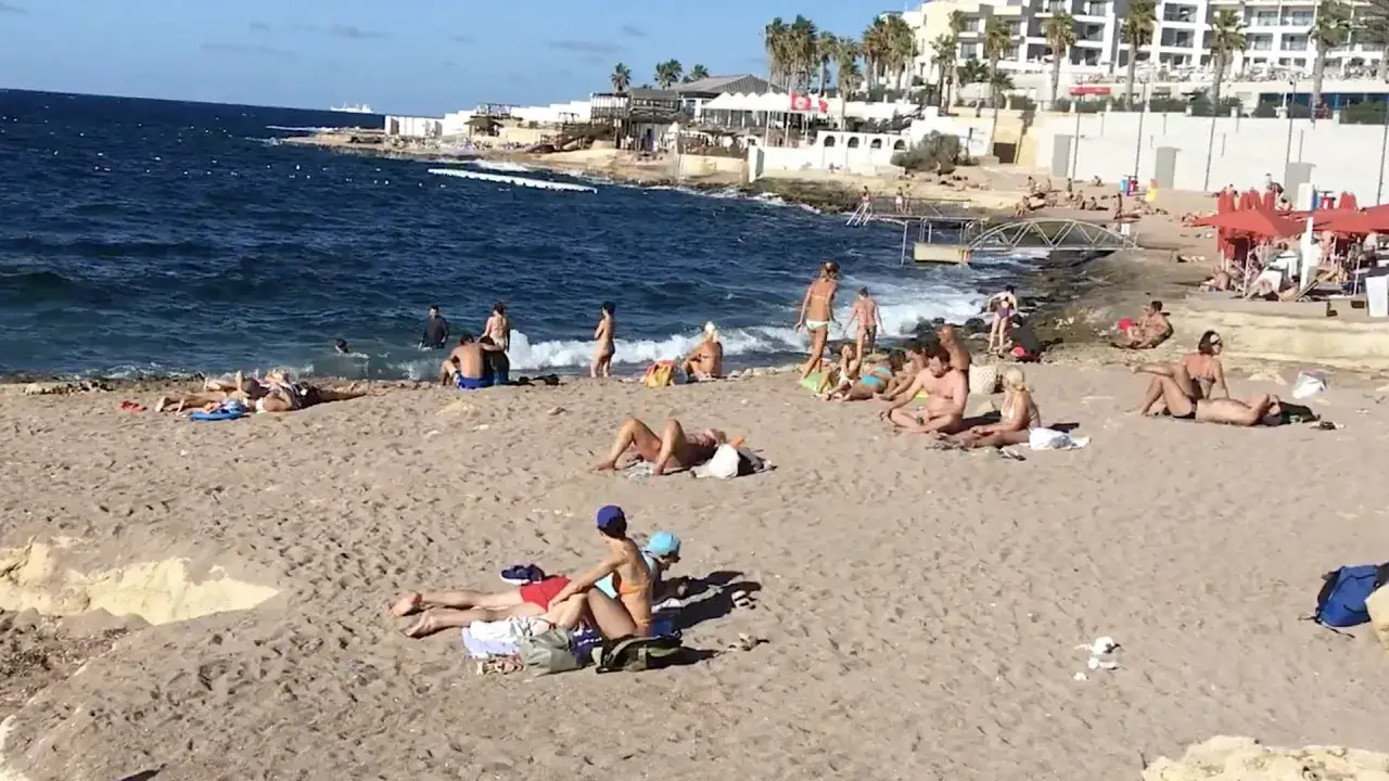 Słoneczny dzień na Bugibba beach. Ludzie opalają się na piasku i kąpią w morzu.
