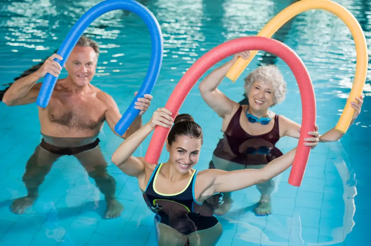 Personas sonriendo y haciendo aqua gym con flotadores de colores en una piscina.