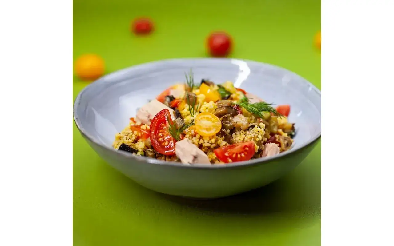 Un piatto colorato di cous cous verdure e tonno, con pomodorini, funghi e aneto, su sfondo verde brillante.