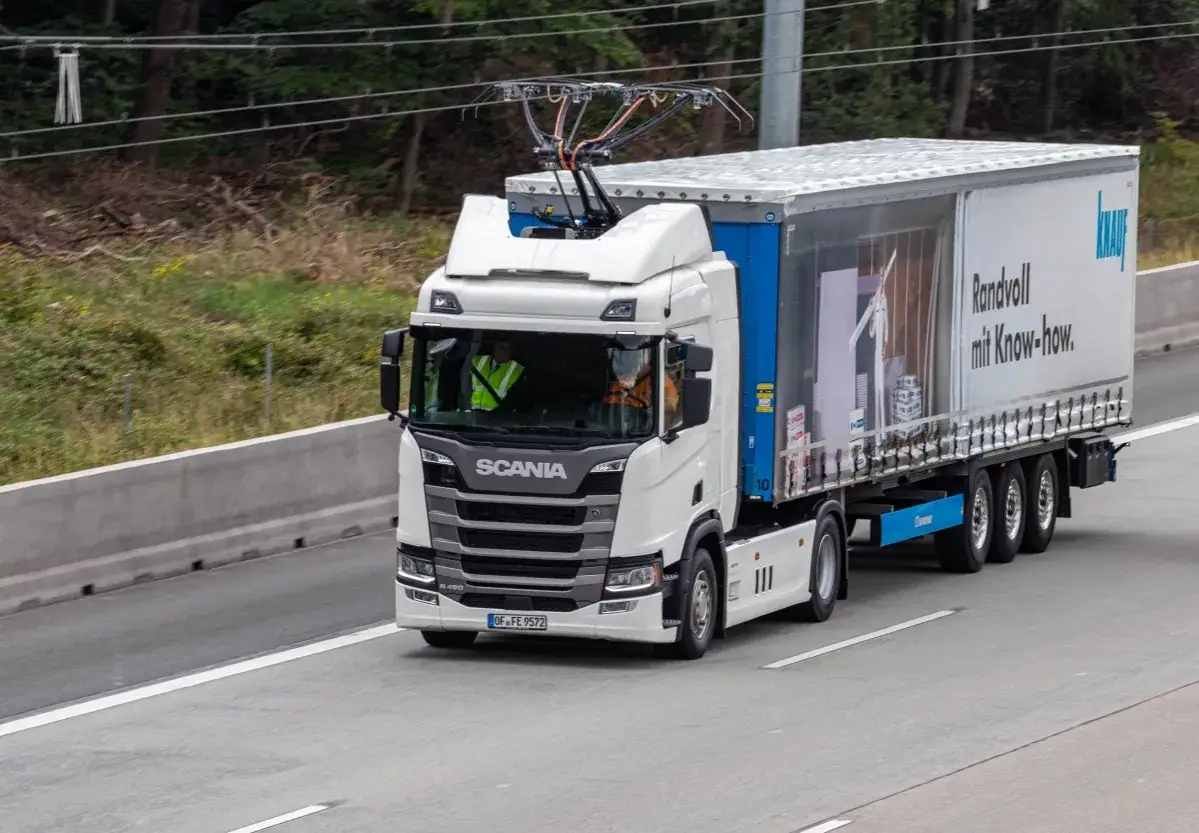 Biała ciężarówka Scania z pantografem na dachu, jadąca po autostradzie. Wygląda jak nowoczesny Peterbilt, gotowy na elektryczną przyszłość.